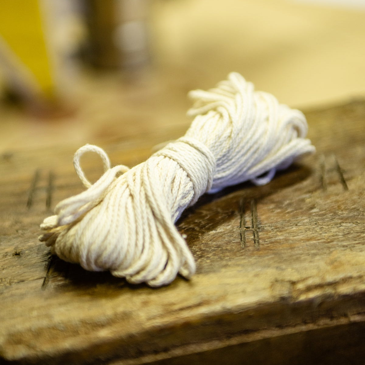 A bundle of off-white cotton wick used for beeswax candle making, resting on a rustic wooden surface with a softly blurred workshop background.