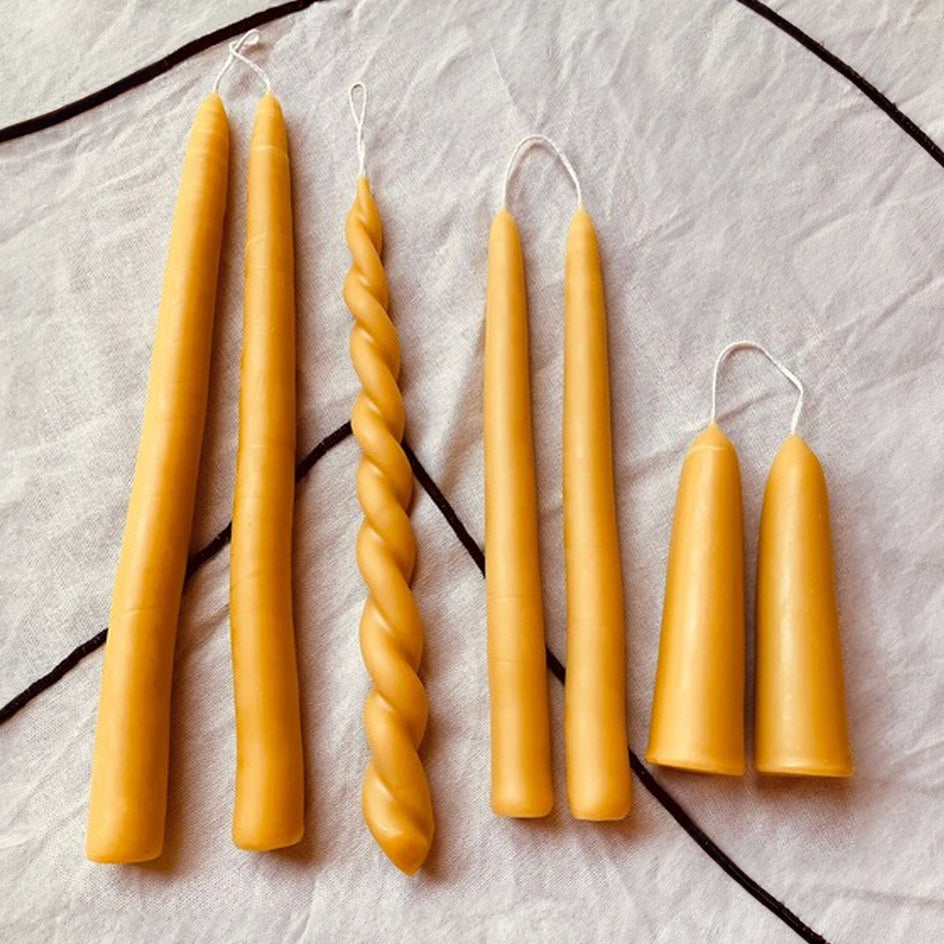 A set of beeswax candles in various sizes with a twisted and braided appearance, displayed on a linen surface with a chain hanging over the edge.