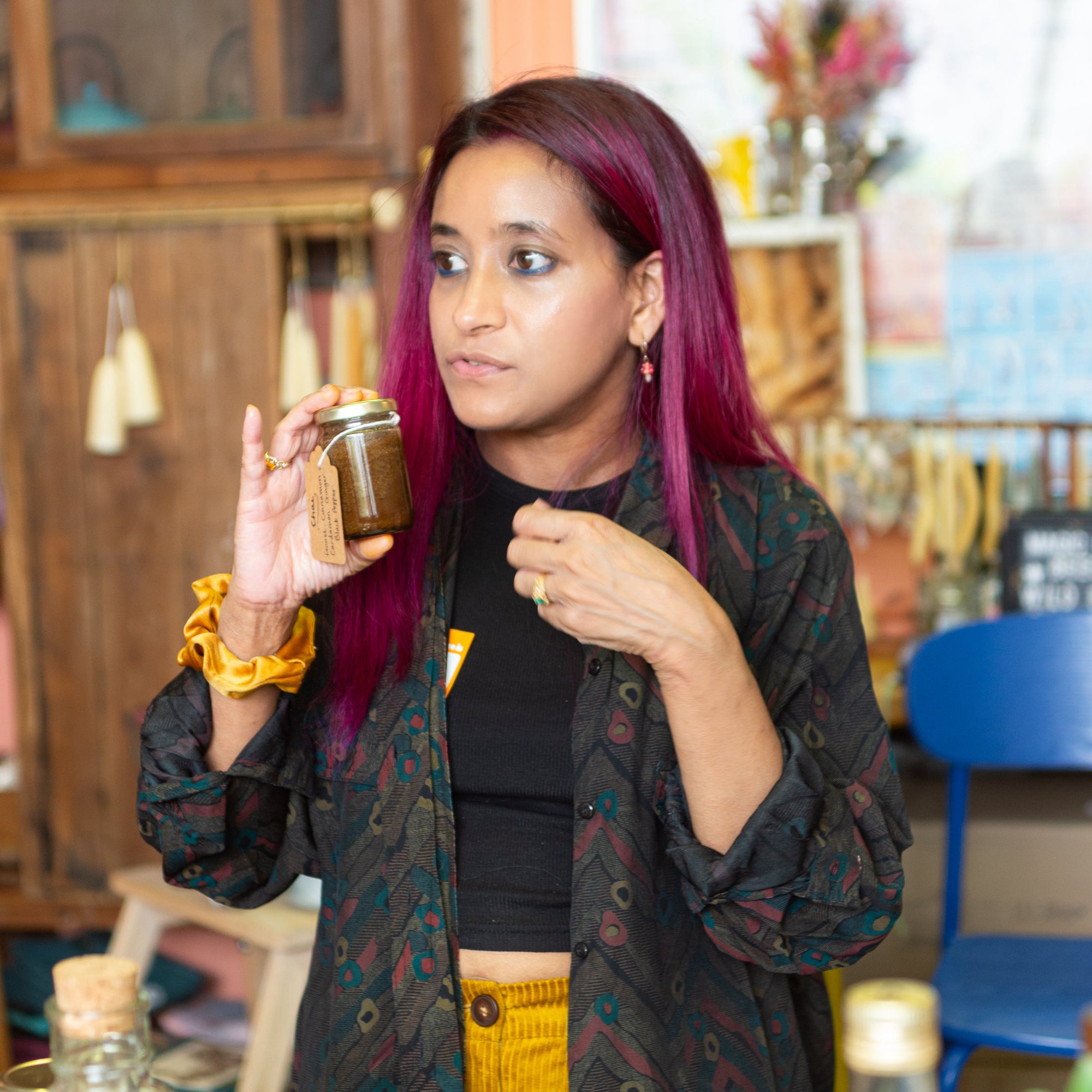Honey, herbs and seasonal botanicals used in our bee-inspired remedies workshop. Moni stands with her vibrant magenta hair in a cozy, rustic work shop holding a small glass jar of honey which is an electuary. She is wearing a black patterned vintage shirt over a black top and yellow cordouroy pants.