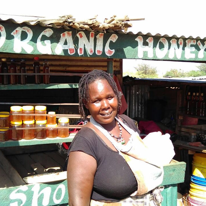 A smiling woman stands in front of a rustic market stall labeled 'ORGANIC HONEY' with multiple glass jars of honey displayed on shelves.