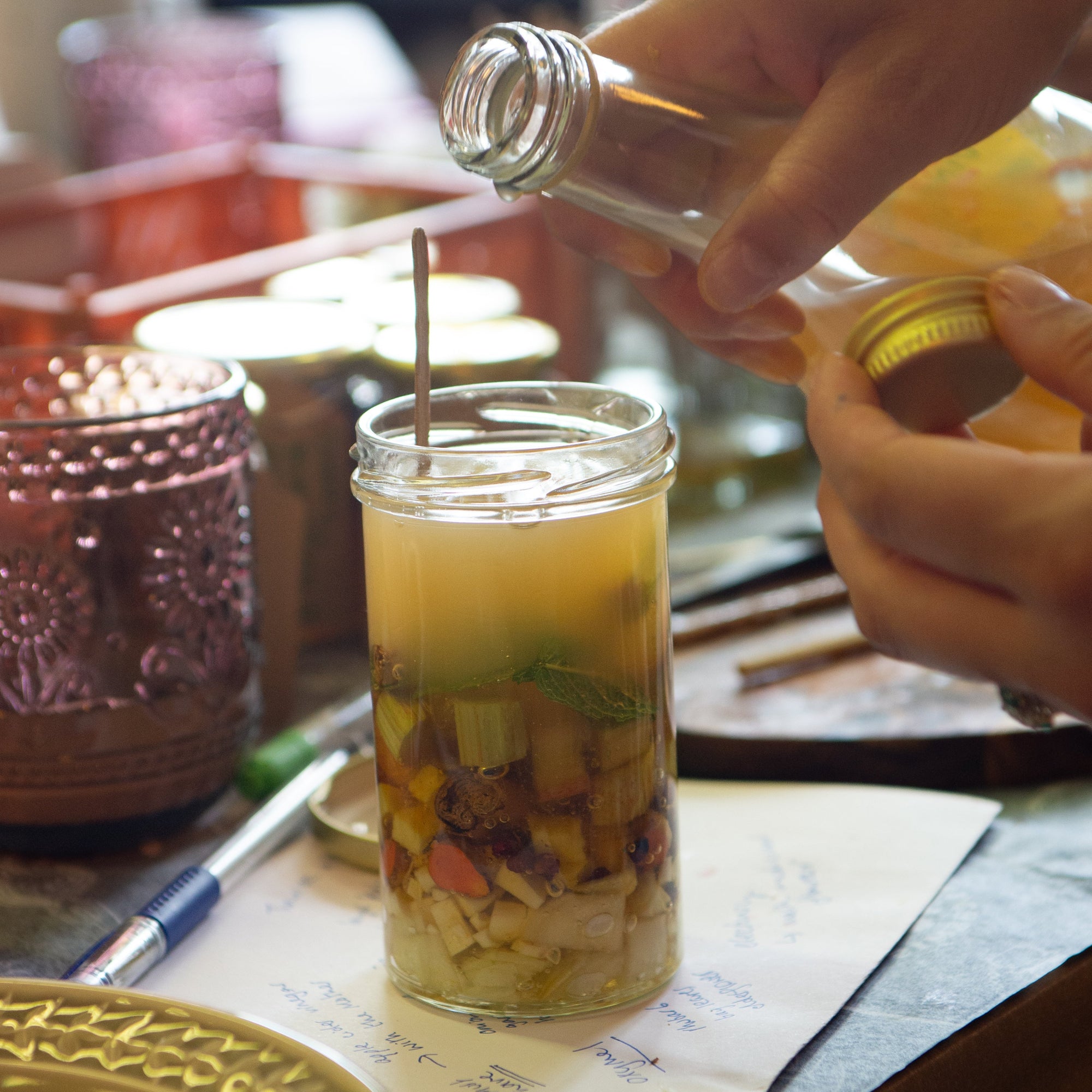 A close-up lifestyle shot of a person pouring a apple cider vinegar from a glass bottle into a clear glass jar containing layered ginger, rhubarb, turmeric, herbs, set on a rustic wooden table with other glassware in a warm, softly blurred background.