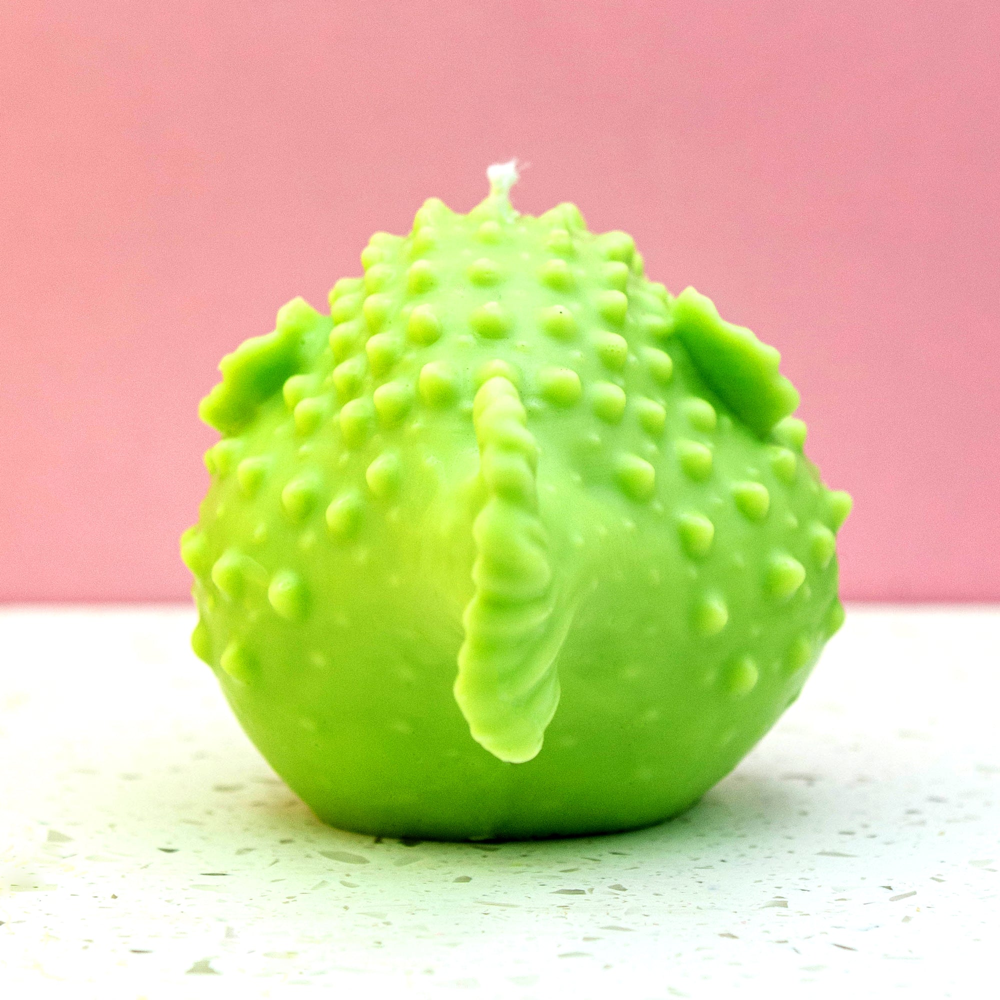 A bright green, textured novelty candle shaped like a spiky lime, set against a pink background and a light speckled surface.
