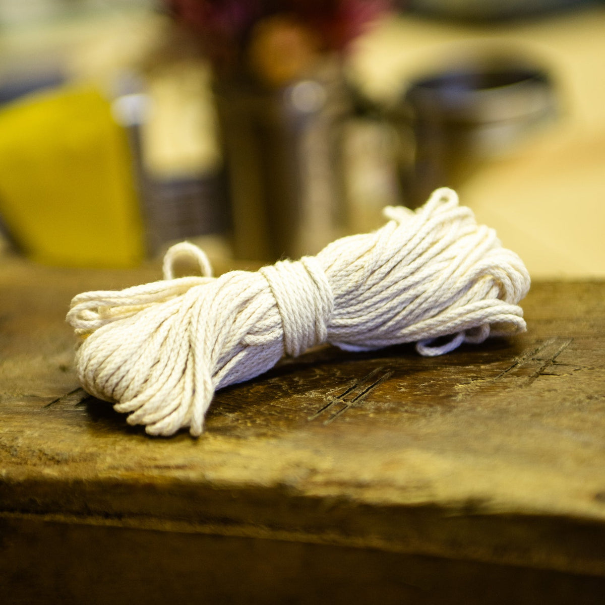 A bundle of off-white cotton cord, twine and wick resting on a rustic wooden surface, with a softly blurred background containing a vase of flowers and other warm-toned objects.