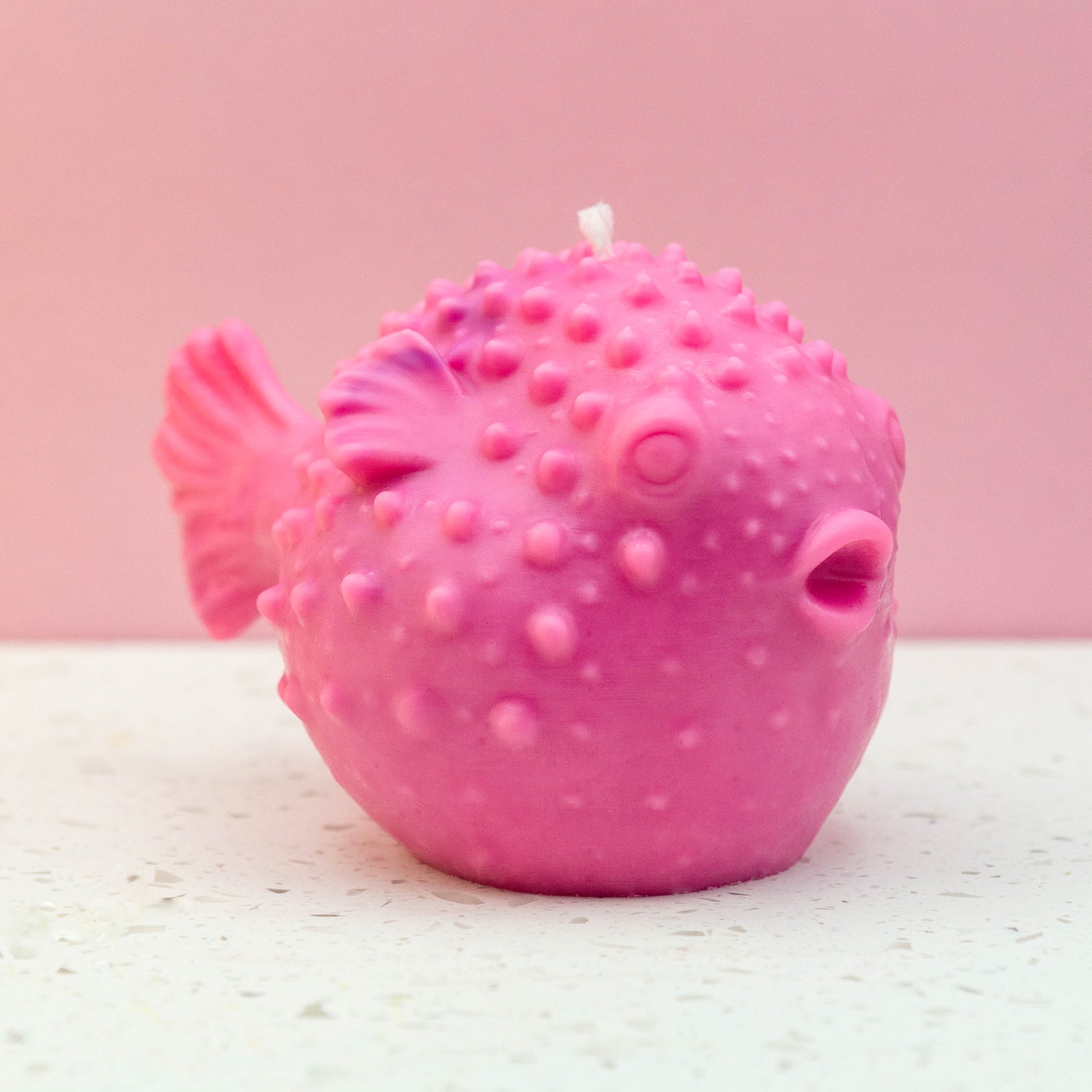 A pink puffer fish shaped candle with a textured, dimpled surface and a small white wick on top, set against a solid pink background and a light speckled surface.