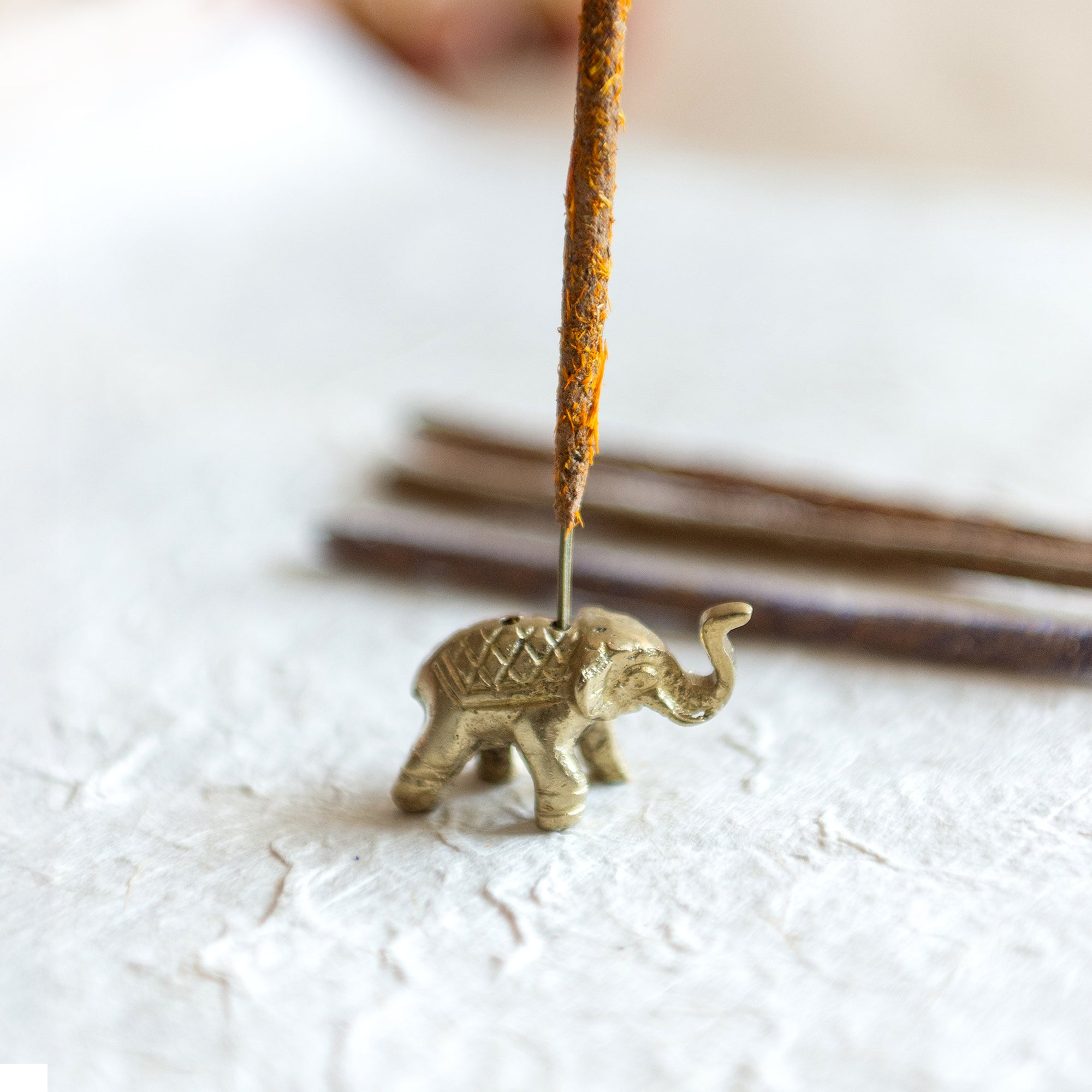 an elephant incense holder, antique gold in colour with 2 holes, holding an incense stick covered in ground calendula flowers which is orange and yellow. the elephant incense holder sits on textured lokta paper with more incense stick faded in the background