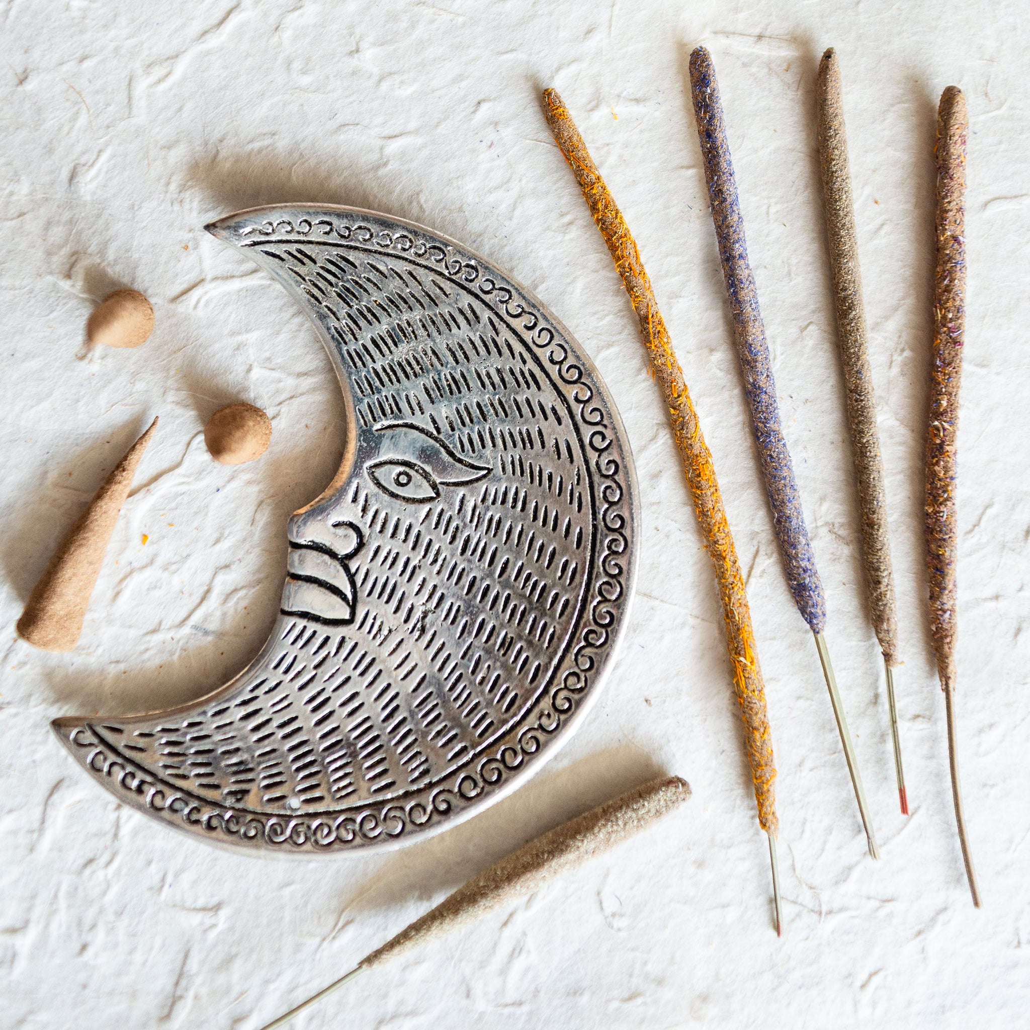 aluminium crescent moon incense holder on a cream lokta paper textured background. the moon incense holder is surrounded by incense sticks that have been handmade in Surrey. There is a calendula incense stick, cornflower incense stick, yarrow incense stick and wildflower incense stick. There also some propolis incense cones, all of which can be made on A beekeepers incense making workshop in Dorking, Surrey.