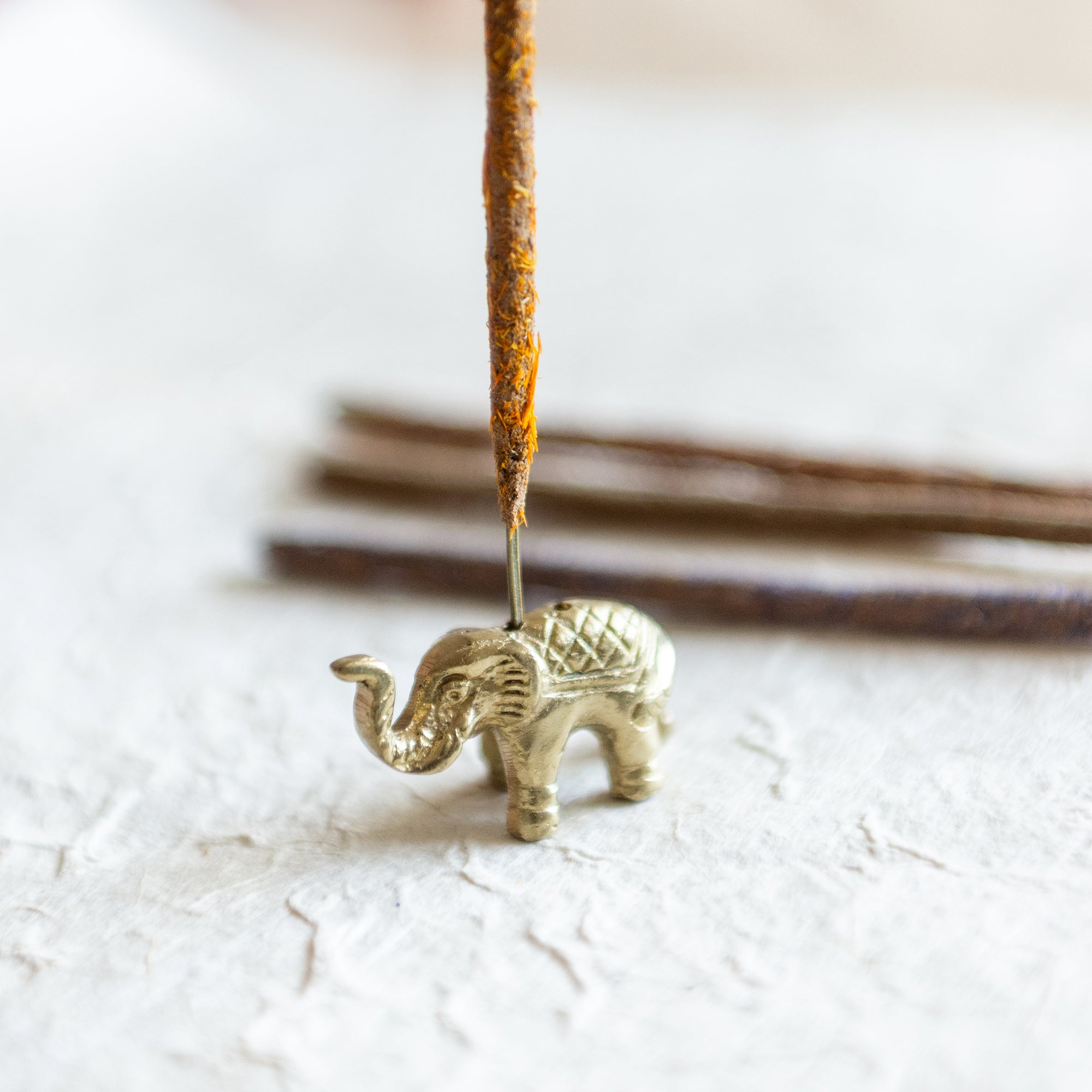 an elephant incense holder, antique gold in colour with 2 holes, holding an incense stick covered in ground calendula flowers which is orange and yellow. the elephant incense holder sits on textured lokta paper with more incense stick faded in the background. The Incense stick has been made from the incense making workshop based in Surrey.