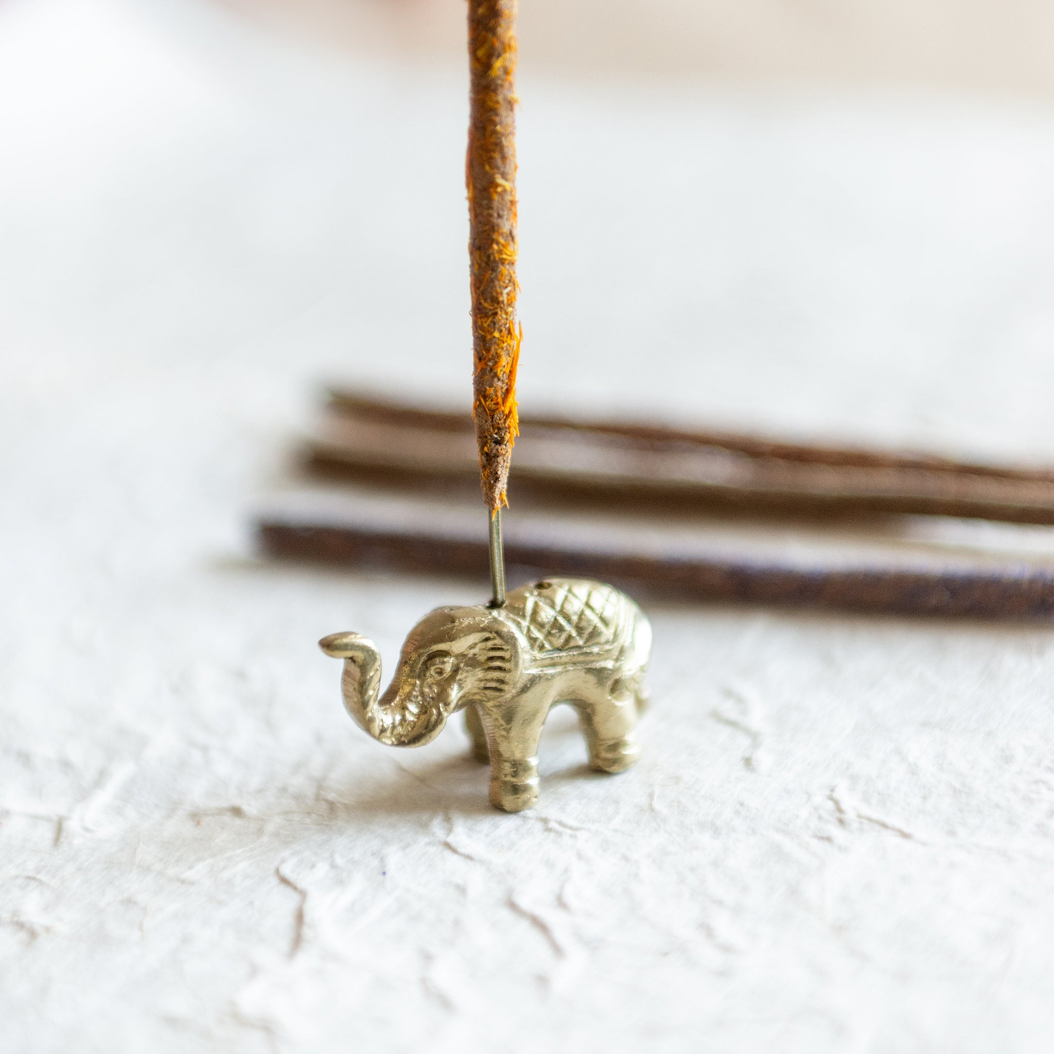 an elephant incense holder, antique gold in colour with 2 holes, holding an incense stick covered in ground calendula flowers which is orange and yellow. the elephant incense holder sits on textured lokta paper with more incense stick faded in the background. The Incense stick has been made from the incense making workshop based in Surrey.