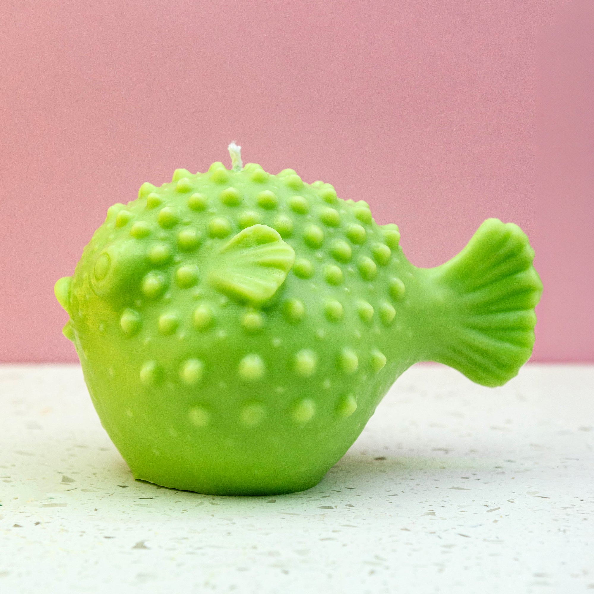 A green puffer fish shaped candle with a textured, bumpy surface and a small white wick on top, set against a pink background and white speckled surface.