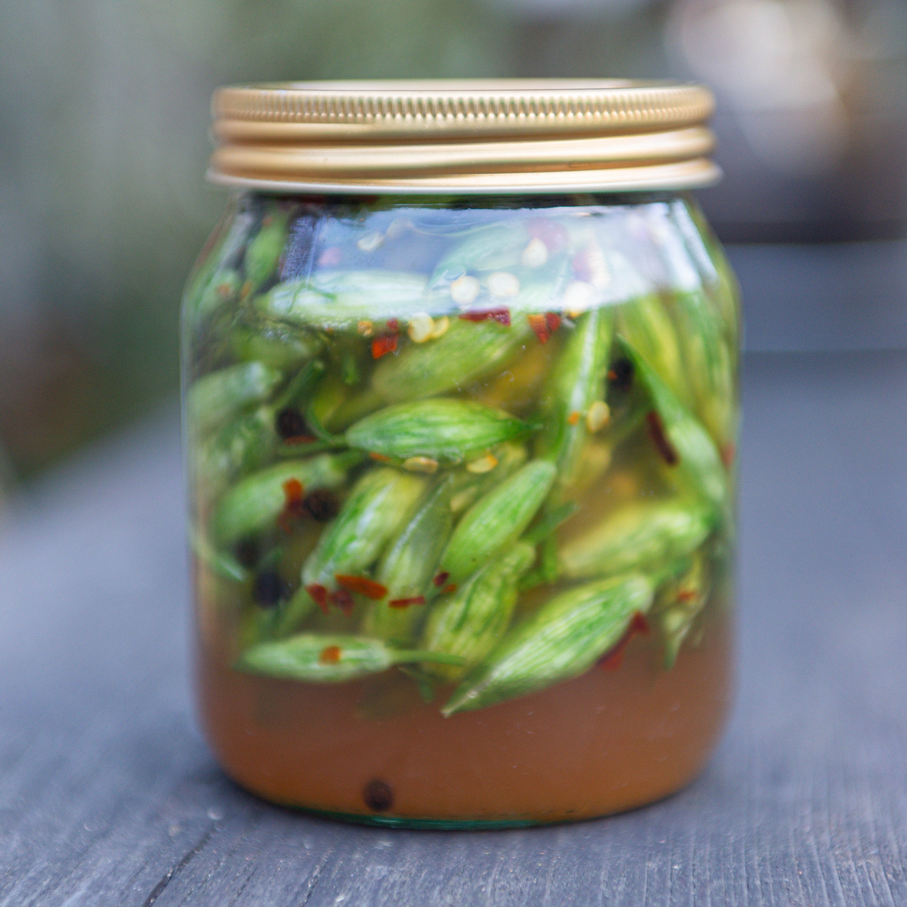 A clear glass mason jar with a gold metal lid, filled with wild garlic buds, preserved and pickled, placed on a wooden surface with a blurred outdoor background.