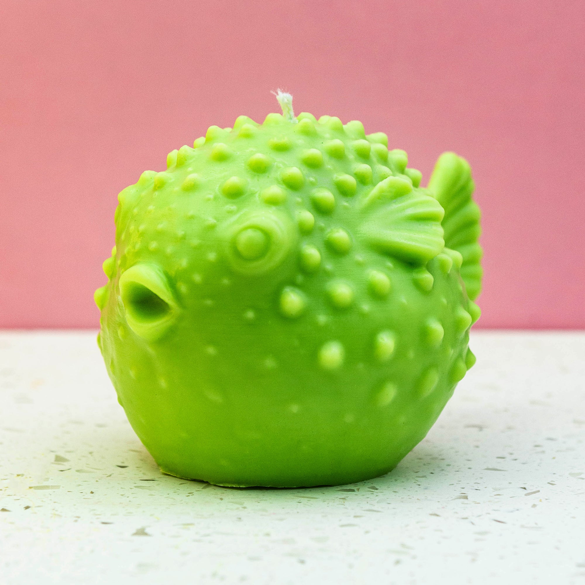A bright green puffer fish shaped candle with a textured, bumpy surface and a white wick on top, set against a pink background and a light speckled surface.