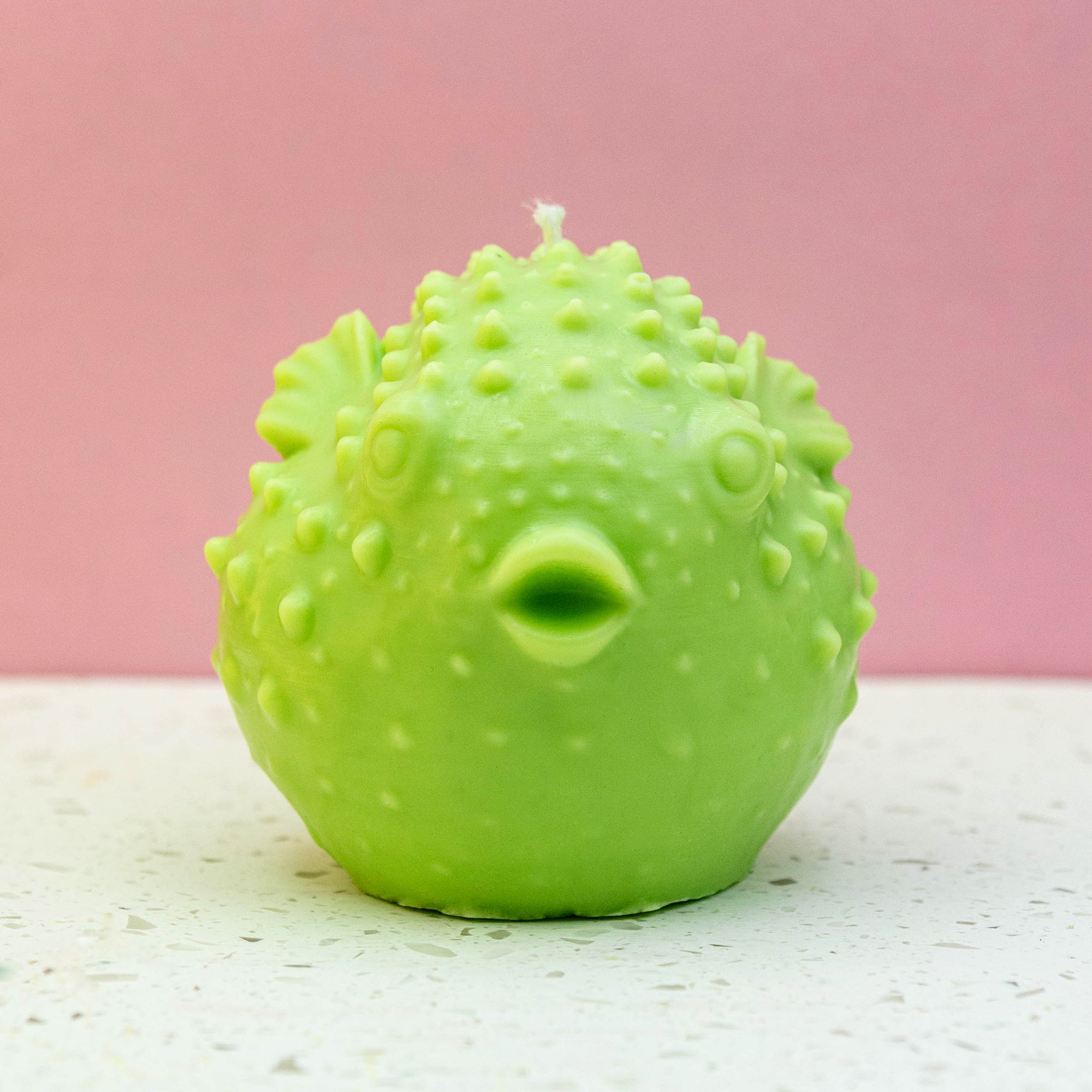 A front facing, bright green puffer fish shaped candle with a textured, bumpy surface and a small white wick on top, set against a pink background and a white speckled surface.  Made in Surrey