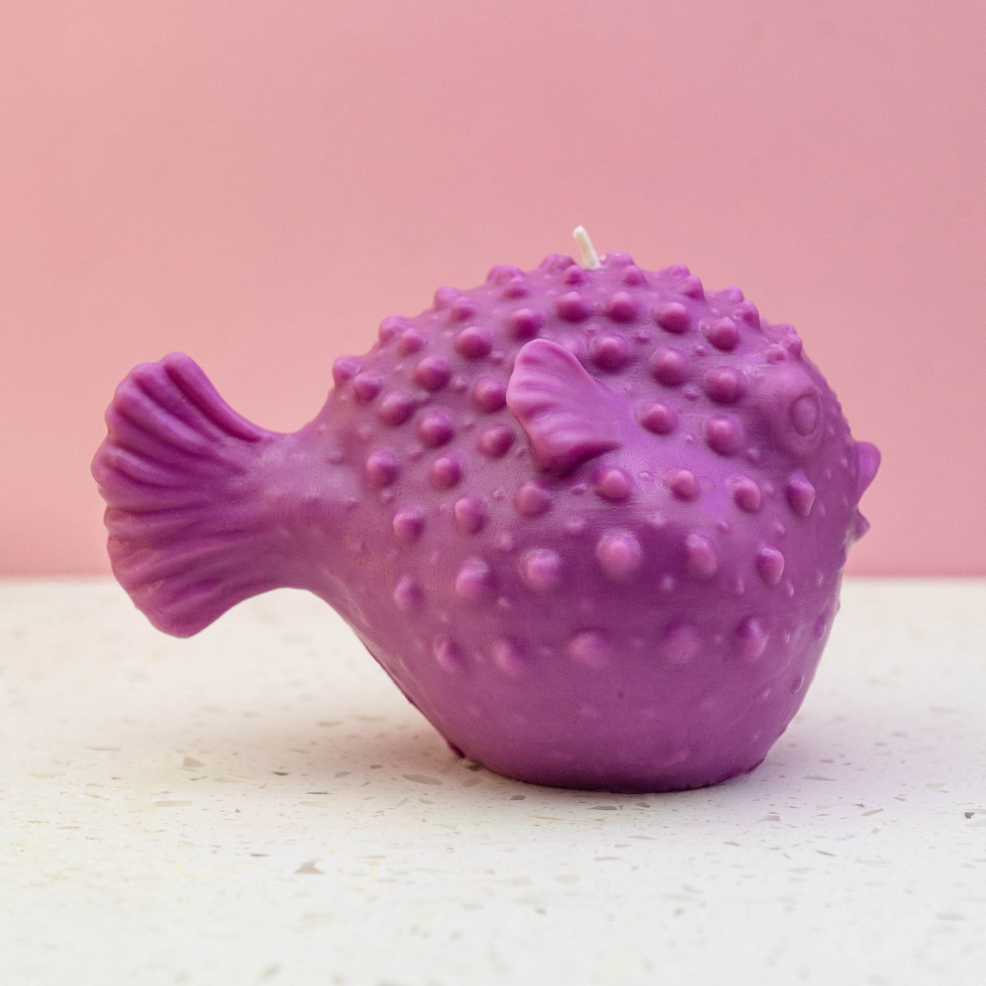A purple puffer fish candle with a textured, bumpy surface and a small white wick on top, set against a soft pink background and a light speckled surface. made in dorking