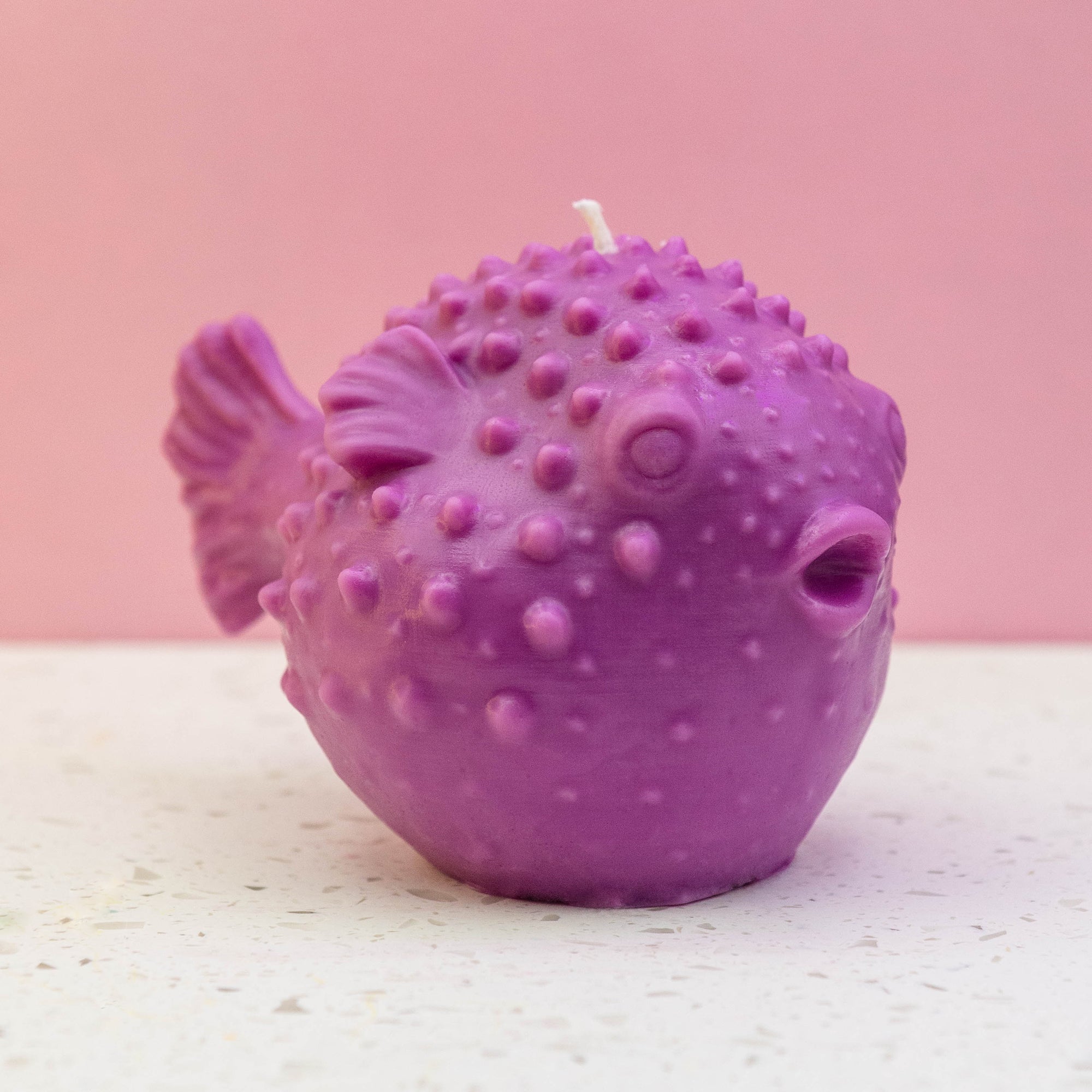 A purple puffer fish candle with a textured, bumpy surface and a white wick on top, set against a pink background and a light speckled surface.