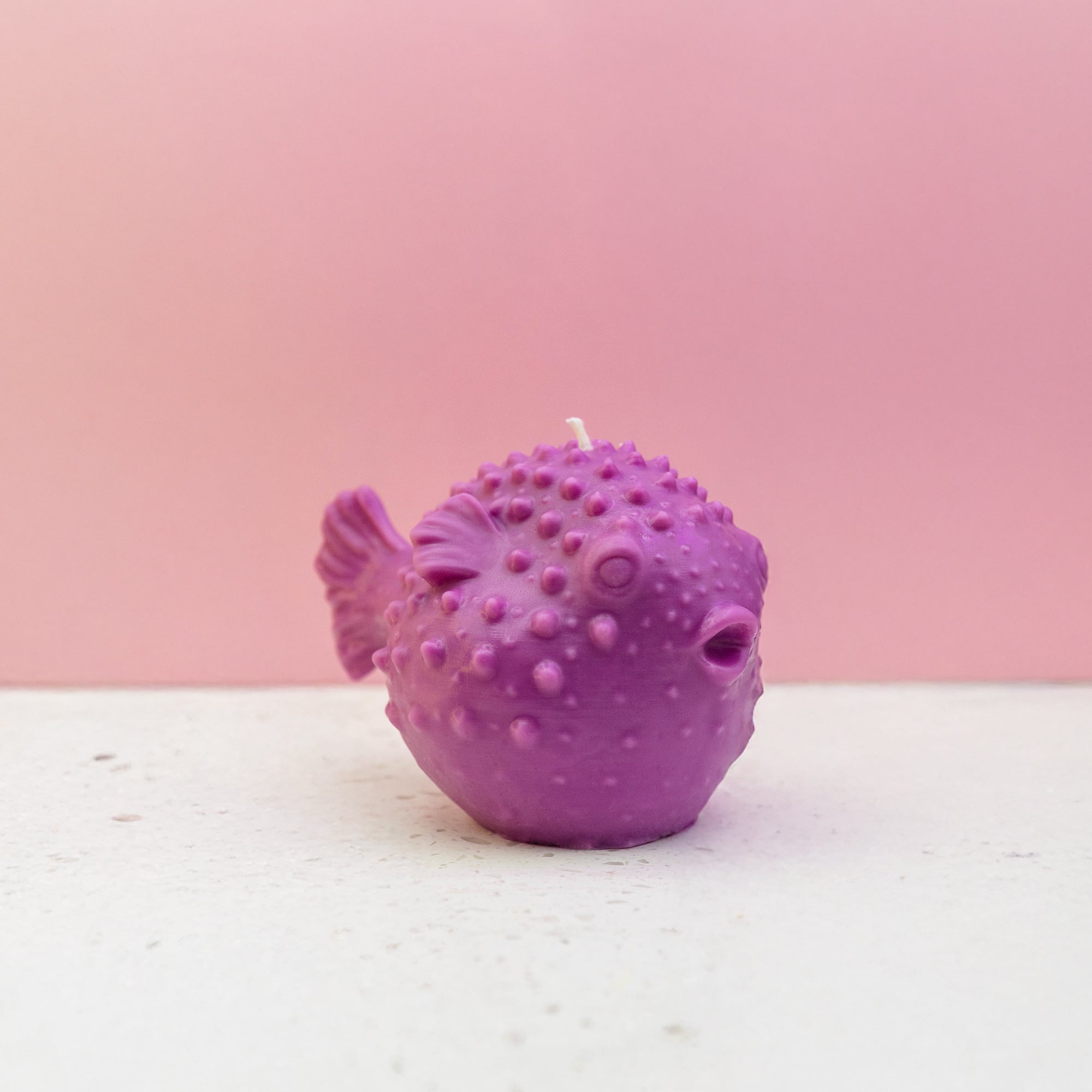 A purple puffer fish beeswax candle with a textured, dimpled surface and a small white wick on top, set against a pink background and a white surface.